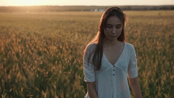 Thoughtful Pretty Girl Is Walking Over Field with Wheat, Sunset Is in Background