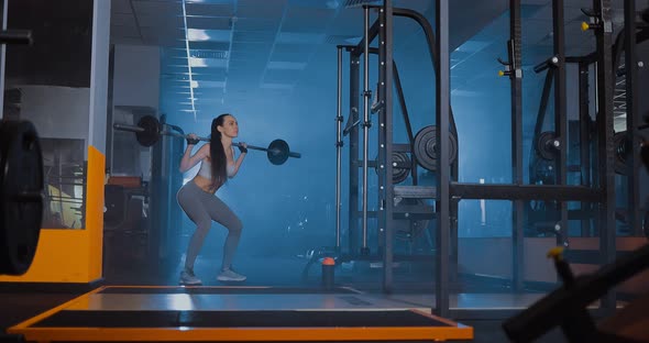 Girl Crouches with a Barbell Behind Her Back in the Gym