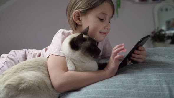 Little girl lying on bed with cat and smartphone