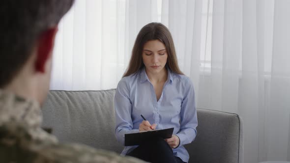 Young Woman Psychologist Filling Anamnesis for Man Soldier Asking Patient and Writing in Notepad at