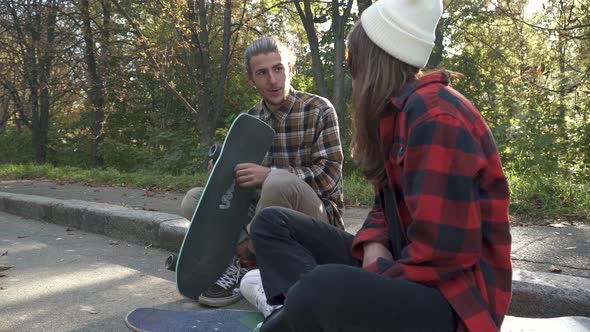 Young Skateboarding Couple Sitting Outdoors. Girl in a Plaid Shirt and White Hat Sitting on a