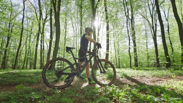 A Cyclist with a Bicycle is Walking Along a Forest Path