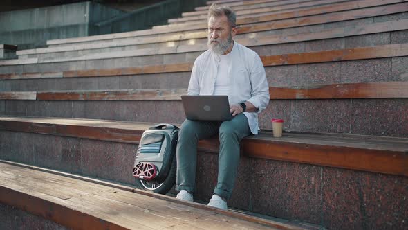 Elderly Man Sits on Stairs Step in Park Working on Laptop