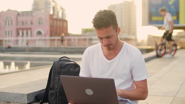 Young Handsome Man in White Tshirt Works with Laptop in the City Center While Sitting on the Steps