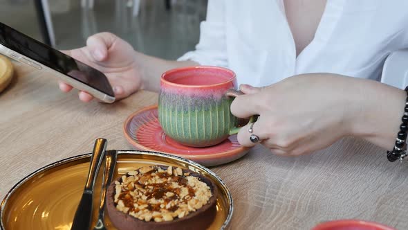 Female Hands in a White Shirt at a Table in a Cafe Holding a Cup of Tea and Using a Smartphone for