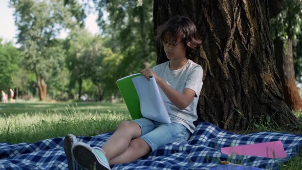 An attractive, curly little boy leaned against a tree and drew something in a notebook