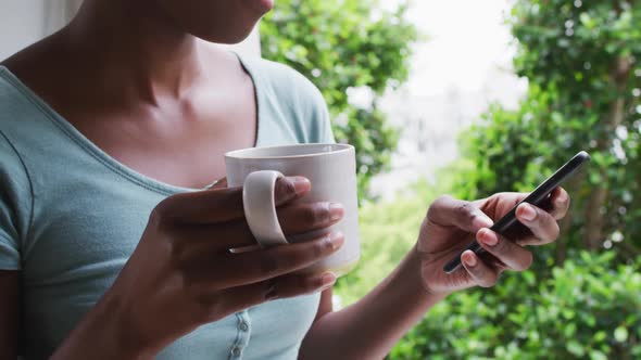 African american woman holding coffee cup using smartphone at home