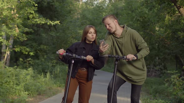 Young Man Shows His Girlfriend Something on Phone They Smile and Drive Away on Their Electric