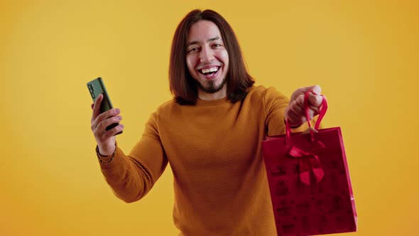 A Long Haired Bearded Happy Young Man Hanging a Phone and a Red Gift Bag in His Right Hand Inside a