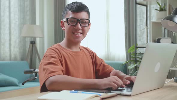 Asian Boy Sitting In A Wheelchair Using Laptop Computer While Looking And Smiling To Camera At Home