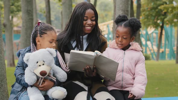 Happy African American Family Resting on Bench in Park Young Mother Reads Book to Daughters Child
