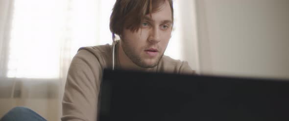 A young man speaking to someone through earphones while working on his laptop. 