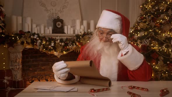 Santa Claus Reading Scroll Wishlist or Letter Sitting at His Desk Near a Christmas Tree and