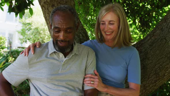 Diverse senior couple in garden taking selfie sitting under a tree and smiling