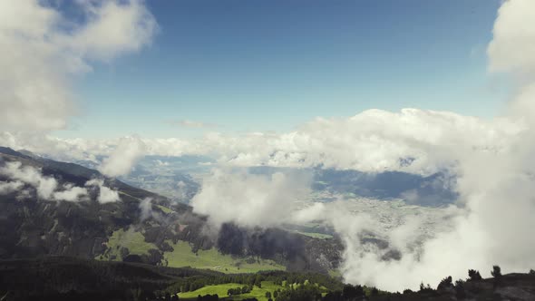 Aerial drone shot of a grass covered mountain top flying through clouds. A valley with a small city,