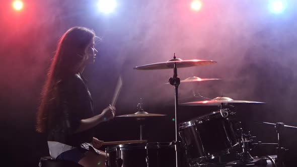 Girl Plays the Drums and Smiles. Black Smoke Background. Red Blue Light From Behind. Side View