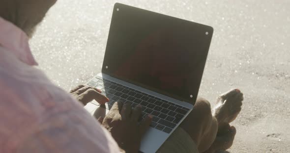 Senior african american man sitting and using laptop on sunny beach