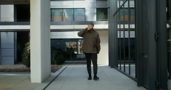 A Young Bearded Man Walks Near an Office Building and Drinks Coffee