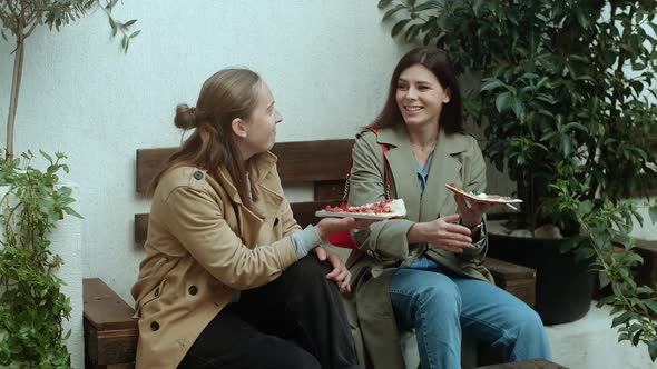 Two young women in coats eating pizza outdoors, having fun together on the terrace in spring time