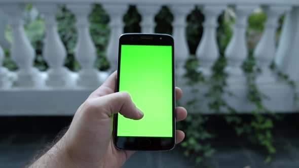 Close up shot of man hand scrolling with smartphone, garden with plants on background. Green chroma