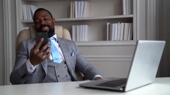 Black Man Is Sitting in Armchair in His Office and Talking By Video Call in Modern Phone