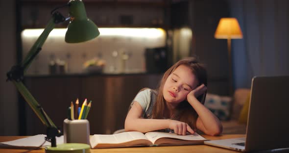 Little Girl Sitting at Table and Reading Book at Home in Evening