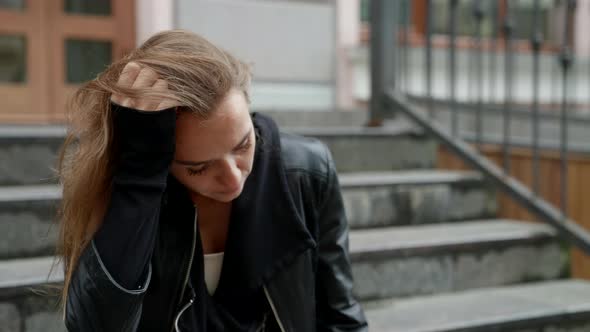 a Sad Blonde in a Black Jacket is Sitting on the Stairs at Home and Leaning Head on Hand