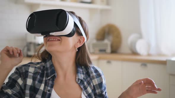 Woman in Virtual Reality Glasses Enjoying Game, Moving Hands and Head Sitting on the Cozy Kitchen