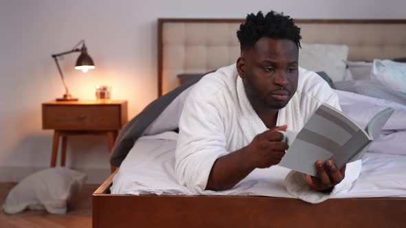 Curios Concentrated Young Man Reading Book Drinking Morning Coffee Lying on Bed at Home