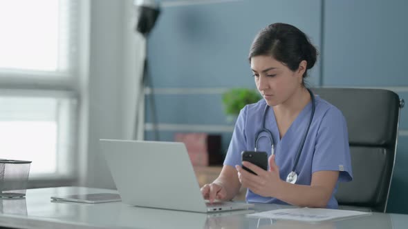 Indian Female Doctor using Smartphone while using Laptop in Office