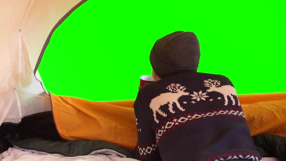 Man in Warm Sweater and Hat is Lying in Open Tent Drinking Hot Tea and Looking at Green Screen