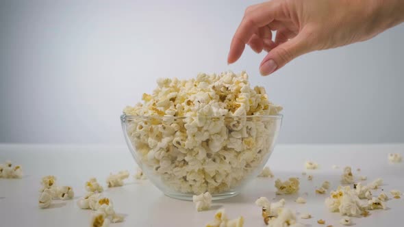 Closeup Woman's Hand Takes Popcorn From Transparent Bowl on White Table Against Background of Light