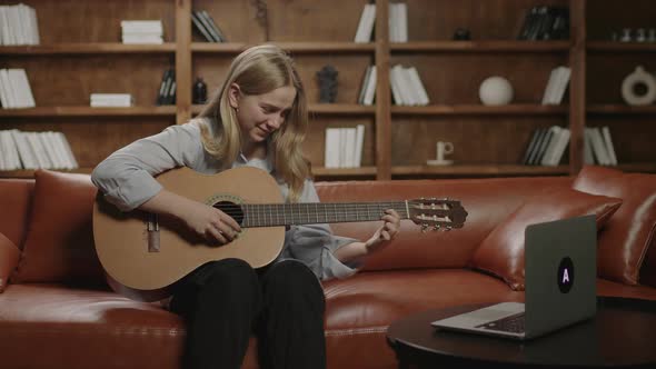 20s Woman Learning To Play the Guitar Looking at Laptop Sitting on the Sofa at Home