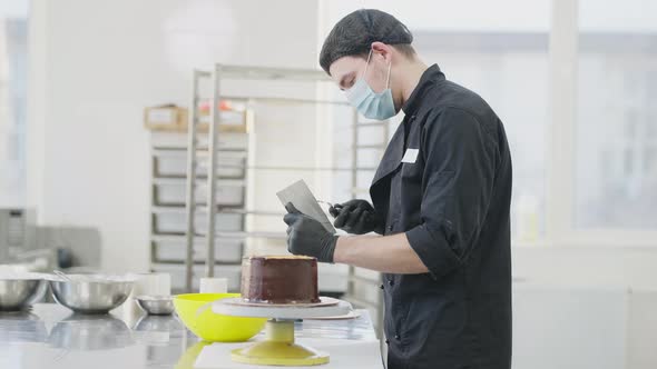 Side View of Professional Focused Man in Covid19 Face Mask Spreading Chocolate on Round Cake on