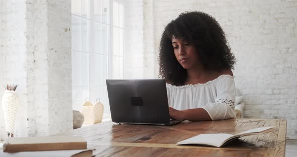 Thoughtful Young Black Woman Freelancer Typing on Laptop Sitting at Table