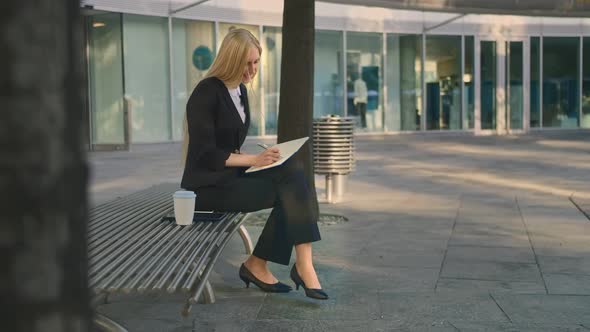 Successful Business Woman Taking Notes Outdoors. Side View of Elegant Girl Sitting on Bench in Patio