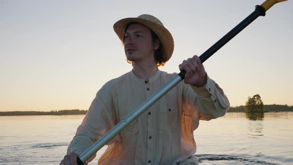 Young Man Does Canoeing on Calm River with Reeds at Sunset