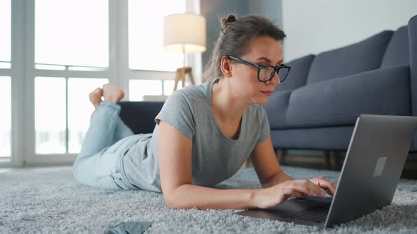 Woman with Glasses Is Lying on the Floor and Working on a Laptop