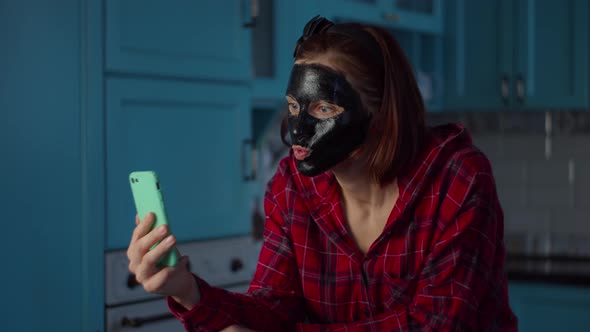 Young 30s woman with black cosmetic mask on her face using mobile phone