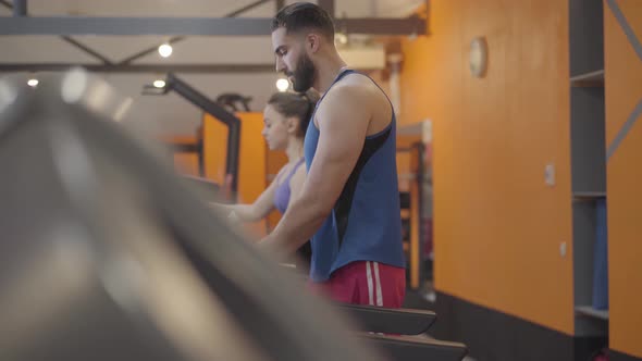 Side View of Middle Eastern Man and Caucasian Woman Training on Treadmill in Gym