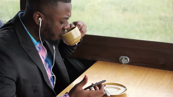 Black Man is Listening Music in Earphones Browsing Smartphone Sitting in Cafe