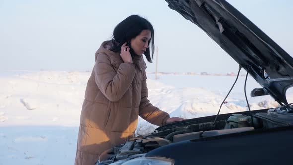 Upset Woman Looks at Broken Auto Engine on Snowy Road