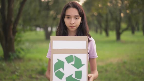 Portrait of Confident Teenage Girl Looking at Camera Standing with Box with Recycling Sign in Park