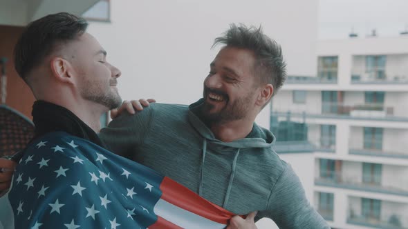 Excited Newlywed Handsome Gay Couple Hugging Under USA Flag on the Balcony