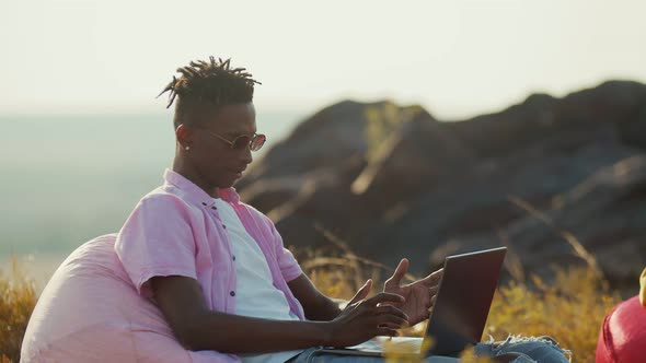 African American Man Sitting with Laptop on Chair Bag in Nature Side View