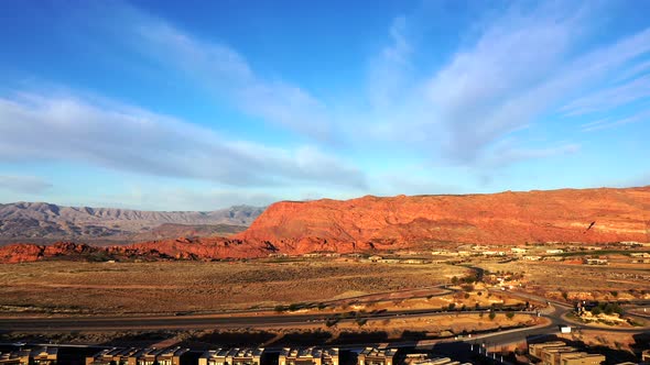 View from a drone of a sunlit city and a road with cars in the foreground and red mountains on the h