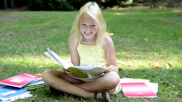 Little Cute Smart Girl Reads a Book in the Park with Many Workbooks Around and Smiles To Camera