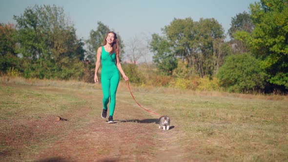 Beautiful athletic woman in sporty turquoise overalls walks with her fluffy cat on a leash in forest