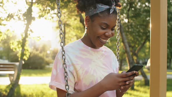 Joyous African Woman Sitting on Swing in Park and Using Phone