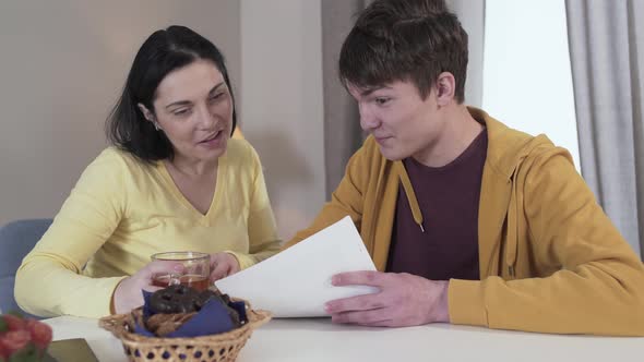 Portrait of Beautiful Brunette Caucasian Woman Drinking Tea As Her Teenage Son Coming Up and Showing